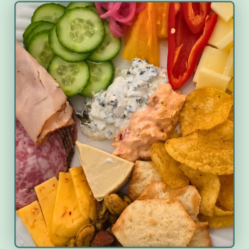 Overhead view of a colorful snack plate with protein, crackers, fresh vegetables, fruit, healthy fats, dips, and a sweet treat, showing an easy balanced no-cook girl dinner.